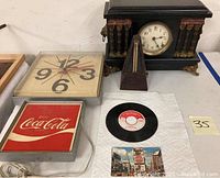 Photo showing Coca Cola illuminated clock, Sessions mantle clock with lion heads, wooden metronome, 45 rpm record, and Coca Cola postcard arranged on table.