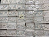 Image showing a collection of 27 rectangular majolica ceramic tiles arranged closely together, showcasing raised floral and geometric patterns in light cream and pastel tones.