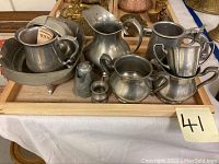 Photo showing assortment of pewter items including bowls, cups, pitchers, creamer, and small containers arranged on a wooden tray.
