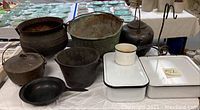 Wide shot showing the collection of cast iron pots, copper kettles, and white enamel kitchenware, arranged on a table with plates in the background.