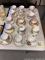 Top angled photo showing 14 porcelain teacups and saucers with diverse floral designs and gold trim, arranged on a white table