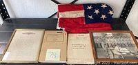 Photo showing all items: folded American flag, framed historical documents, limp book titled Maintenance and Care of Hand Tools, and framed black and white photo.