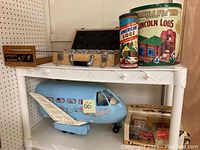 Shelf showing a large blue Barbie airplane playset, the Shoot to the Moon wooden game, and tall tins of Lincoln Logs and American Logs wooden building sets.