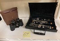 Photo showing the open black case containing the two-piece black clarinet with silver keys and the binoculars in their brown leather case placed beside it.