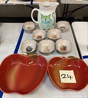 Photo showing white ceramic pitcher with green cat design and seven stoneware fruit baking molds along with two red apple-shaped ceramic bowls.