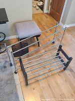 Photo showing one metal shoe rack in front with a padded grey footstool and other metal shoe rack behind it.