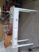 Side angle of white console table with glass top showing drawer, tapered legs, and scratches/scuff marks. Small items and lamp on top.