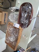 Photo showing various clear glass drinkware arranged in two wicker baskets along with a metal cocktail shaker and glass lidded dish.