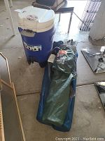 Blue and white Igloo Sport 5-gallon cooler with spigot, shown next to three folded outdoor chairs in carrying bags.