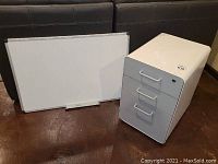 Whiteboard and filing cabinet shown together, whiteboard leaning against couch, cabinet next to it on floor.