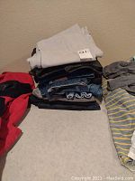 Stack of ten men's jeans folded on a surface showing various colors and fabrics including blue and light gray.