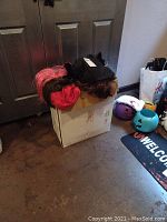 Cardboard box with Halloween wigs in pink, black, and brown colors on top and plastic pumpkin buckets nearby.