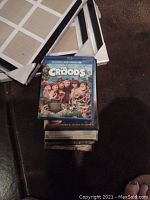 Eight DVDs stacked on the floor with "The Croods" on top, accompanied by white multi photo frames in the background.
