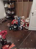 Photo of miscellaneous Christmas decorations including two electric Santa and Mrs. Claus figurines, blue ornaments in a tall tube, a penguin figurine, Christmas lights, and red glitter decorations.