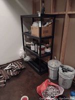 Front view of black plastic shelving unit placed in basement corner, showing four shelves with some storage items on them (not included).