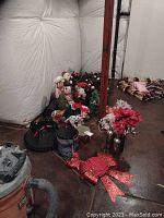 Photo showing metal Christmas tree stand, standing Santa and snowmen figurines, poinsettia garland, glittery red bow, and various smaller holiday items on the floor in basement.