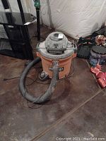 Orange Ridgid wet dry vacuum cleaner with grey top, hose attached, unit placed on concrete floor with other household items near it.