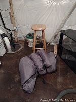 Photo showing wooden stool standing upright with a gray sleeping bag folded on the floor nearby in a basement setting.