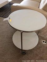 Round glass and metal sofa/corner table with metal legs and black caster wheels on carpet, top and lower shelf visible.