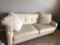 White leather sofa with two fluffy throw pillows on a carpeted floor, showing full front view.