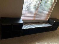 Three black shelf units arranged together under a window, two smaller cube units flanking a larger unit with a gray and white chevron patterned cushion on top.