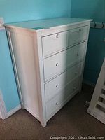 Front view of the white painted wood 5-drawer dresser next to a crib in a room with light blue walls.