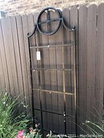 Full view of the metal garden trellis leaning against a wooden fence with flowers at the base.