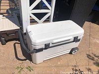 White Igloo rolling cooler with black telescoping handle and wheels on outdoor ground.