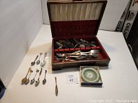 Wide shot of all items including a brown vintage case containing forks and spoons, souvenir spoons laid out, and green decorative plate in a box.