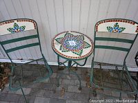 Full view of the 3 piece bistro set showing round mosaic tile table and two matching folding chairs with mosaic tile floral inlays on backrests