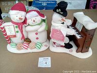 Pair of snowmen figurines and a snowman playing piano on cardboard surface showing Hallmark tags and MaxSold auction card.