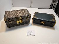 Two closed jewelry boxes shown side by side on a white surface; one floral patterned metal box with gold hardware, one green box with gold trim.