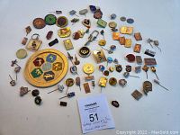 Wide assortment of vintage pins laid out on white surface showing a variety of shapes, colors, and designs honoring Olympics and sporting events from 1974 to 1988.