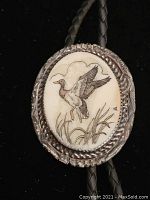 Front view of the bolo tie slide showing a detailed oval silver frame with an engraved duck flying above reeds on a cream background, attached to black braided leather cords.
