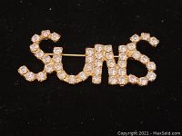 Close-up image of the brooch showing the word 'Suns' spelled out in clear rhinestones with the pin attachment visible at the back.