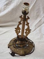 Front and angled views of vintage metal table lamp base with ornate decorative detailing and electrical cord wrapped around base.
