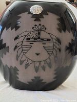 Front view of black and white ceramic vase showing central Kachina face and feather headdress with geometric pattern background.