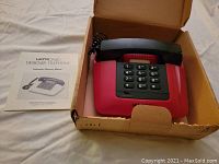 Red Ultronic Designer Telephone with black handset, keypad showing numeric and function keys, in its original box with instruction manual.