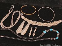 Nine pieces of assorted jewelry displayed on black surface including silver-tone braided necklace, two silver-tone bracelets, two pairs of silver-tone earrings, and two dark brown beaded necklaces with turquoise accents.