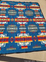 Overall view of large, colorful Pendleton wool blanket with blue background featuring Native American-inspired geometric designs in red, orange, yellow, white, and brown.