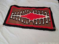 Full view of rectangular Southwest style rug laid flat on light fabric, showing central geometric red and white pattern with brown and black background.