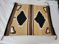 Full view of the rectangular woven rug laid flat, showing symmetrical geometric pattern with two black diamond shapes framed by white and tan, surrounded by stripes of brown, tan and black.