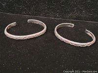 Pair of sterling silver cuff bracelets with engraved floral vine patterns displayed on a black background.