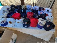 Display of 25 hats arranged in rows on a table including various sports team caps, event caps, and branded caps in multiple colors