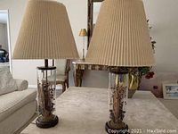 Two table lamps with beige pleated shades and glass cylindrical bases containing dried floral arrangements, shown on a table in a living room setting.