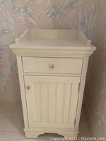 Photo of small cream-colored dresser with one drawer and one cabinet door with vertical panel details and crystal knobs