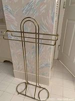 Freestanding gold-tone vertical towel stand placed against patterned wallpaper near door, showing overall design and condition with scratches