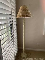 Tall floor lamp placed in a corner beside a window with white blinds, showing a woven basket-style lampshade and a twisted carved pole with round base.