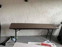 Photo showing a long rectangular foldable table with a wooden top and foldable metal legs standing against a wall.
