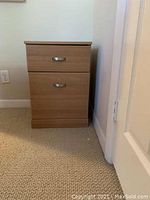 File cabinet with two drawers, wood finish, metal handles, placed on light brown carpeted floor near cream-colored wall